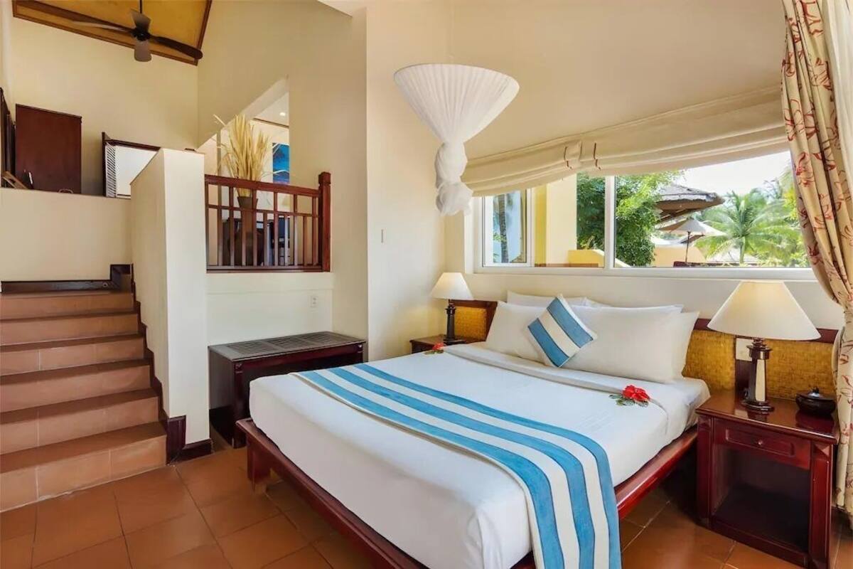 Une chambre d’hôtel avec un grand lit, un escalier, une fenêtre avec des rideaux et une vue sur des palmiers.