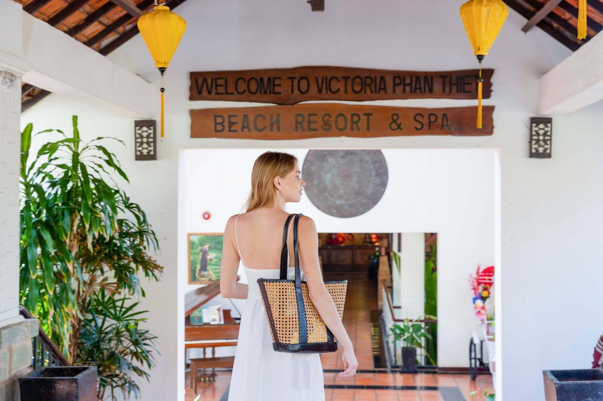 A woman in a white dress standing in front of a wooden sign that reads 'Welcome to Victoria Phan Thiet Beach Resort & Spa.'