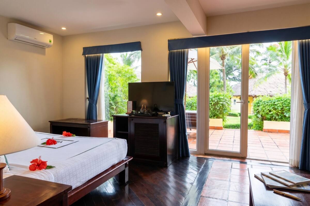 Habitación de hotel con cama, escritorio, televisión y vista a un jardín a través de la puerta de vidrio.