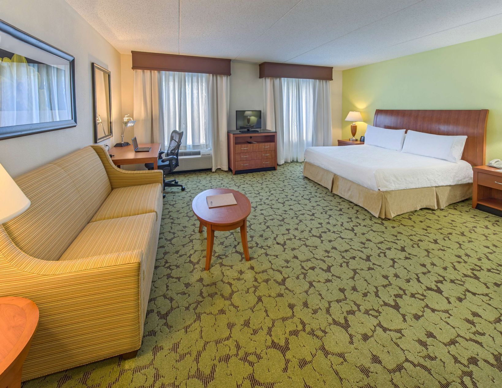 A hotel room with a bed, a sofa, a desk with a computer, a TV, and a small table.