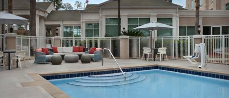 A swimming pool with a white fence, a seating area with cushions, and a building in the background.
