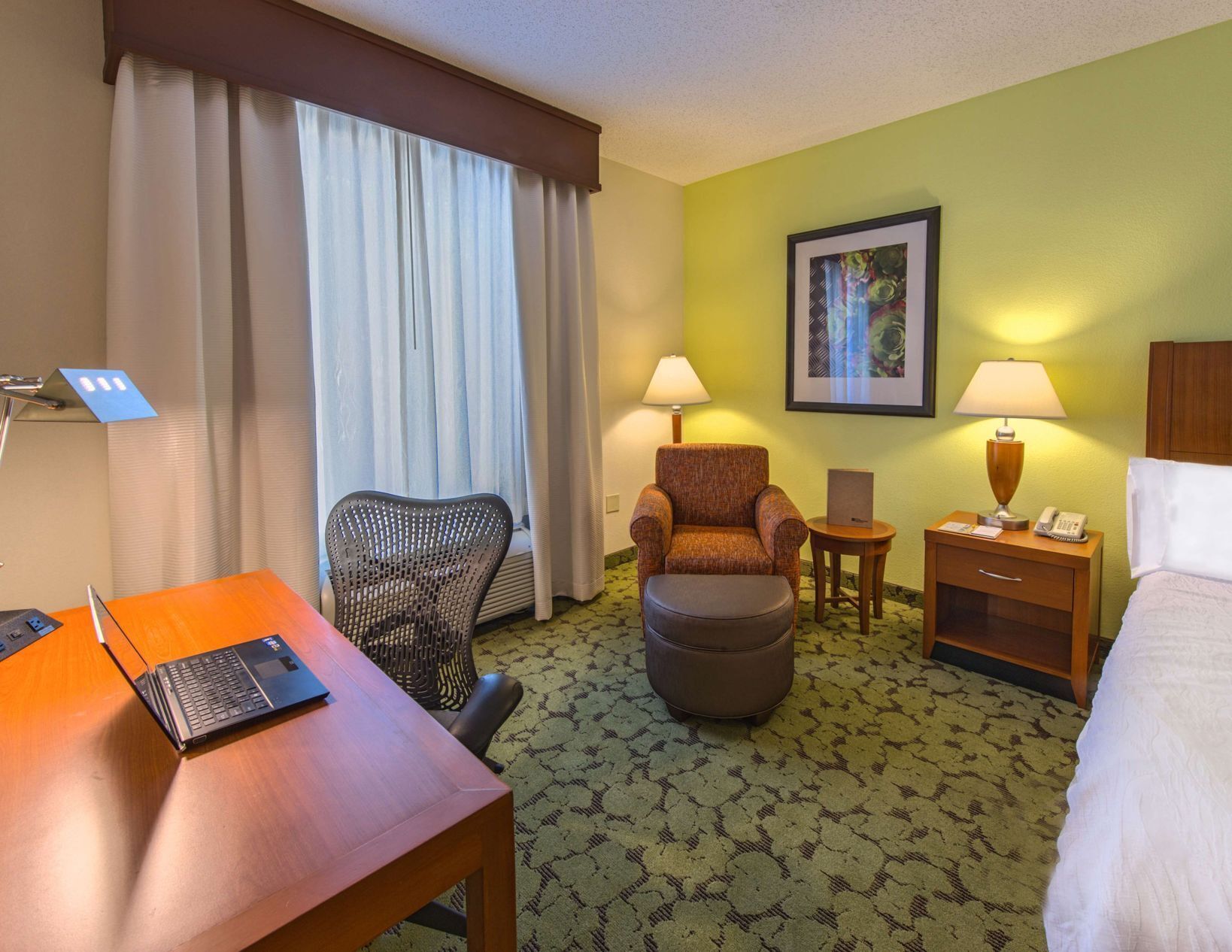 A hotel room with a bed, a desk with a laptop, a chair, a small table, a nightstand with a lamp, and a framed picture on the wall.