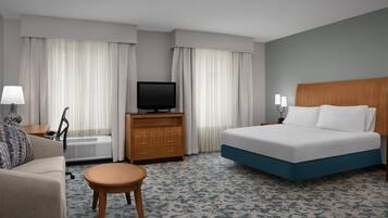 Quarto de hotel com uma cama, televisão, sofá, mesa pequena e janela com cortinas.