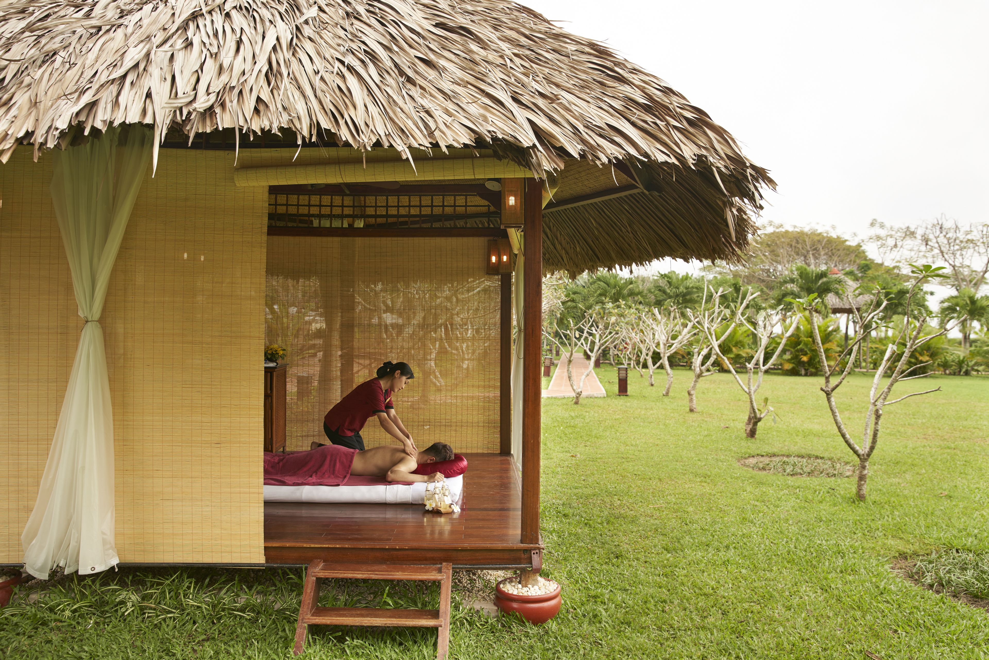 A traditional thatched-roof structure with a wooden bench and a mat, likely used for relaxation or massage, set in a grassy area with trees.