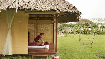 A traditional thatched-roof structure with a wooden bench and a mat, likely used for relaxation or massage, set in a grassy area with trees.