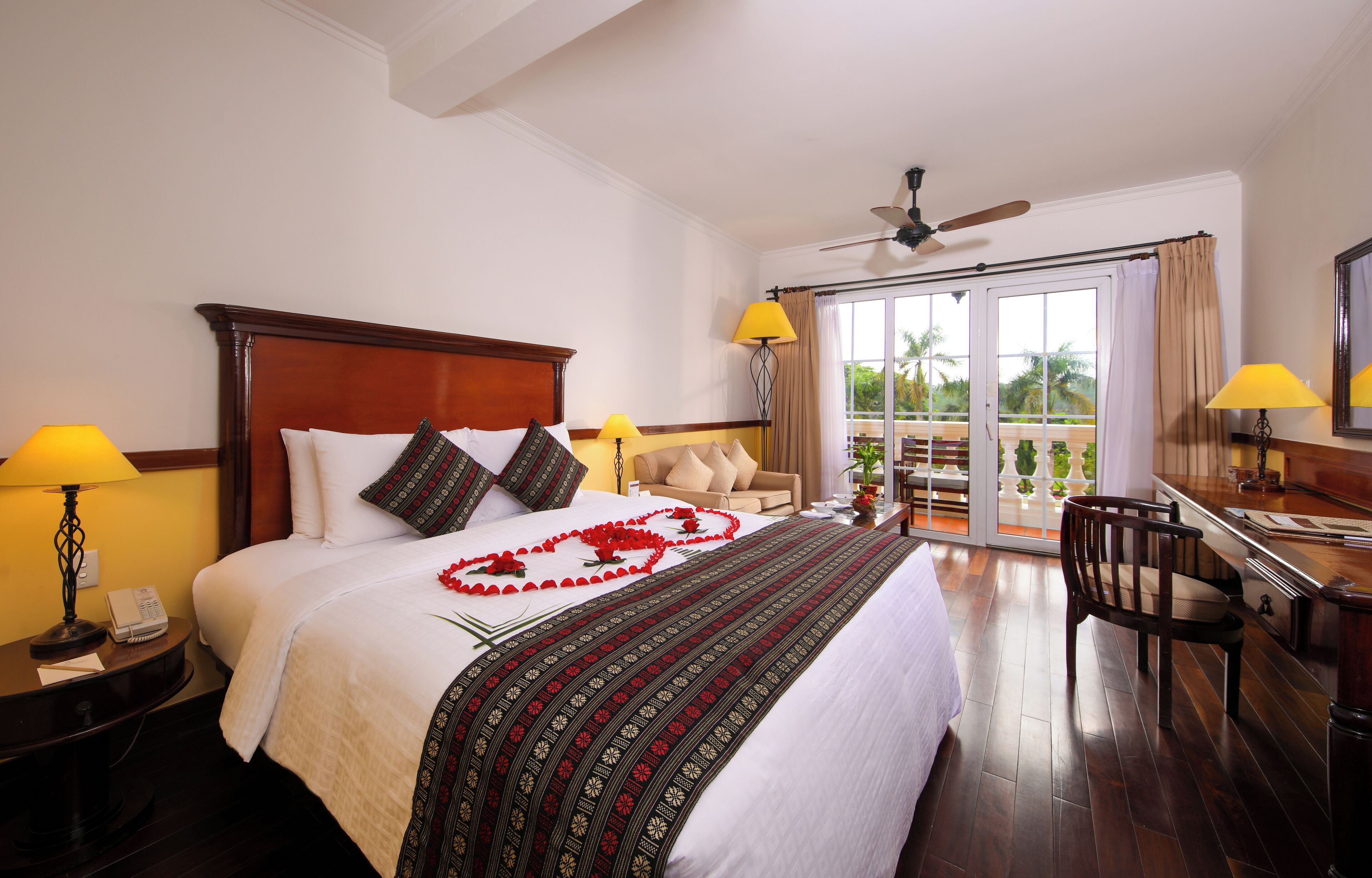 A hotel room with a large bed, a desk, a chair, a balcony with a view, and a ceiling fan.