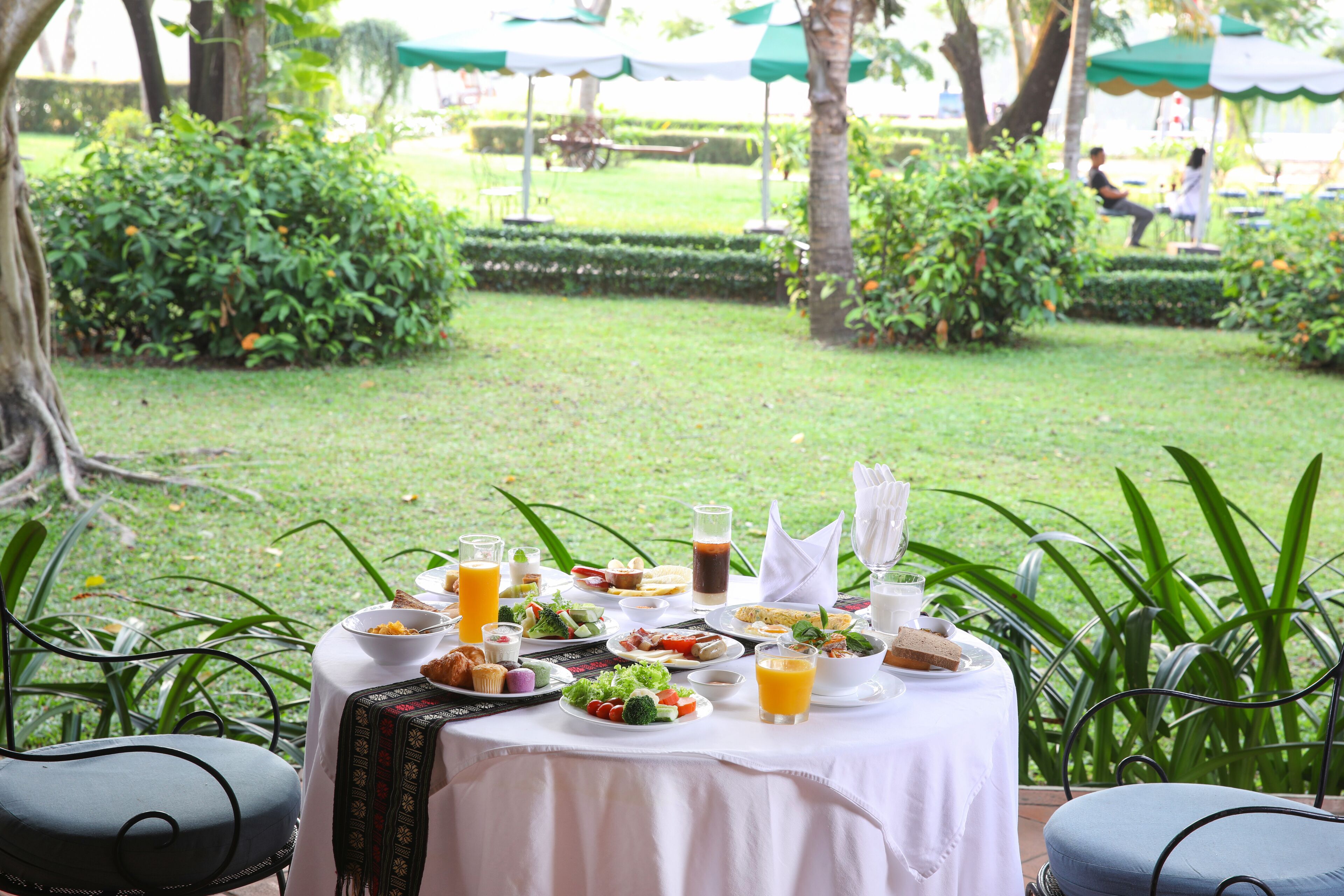 An outdoor dining area with a table set for a meal, surrounded by greenery and a view of a well-maintained garden.