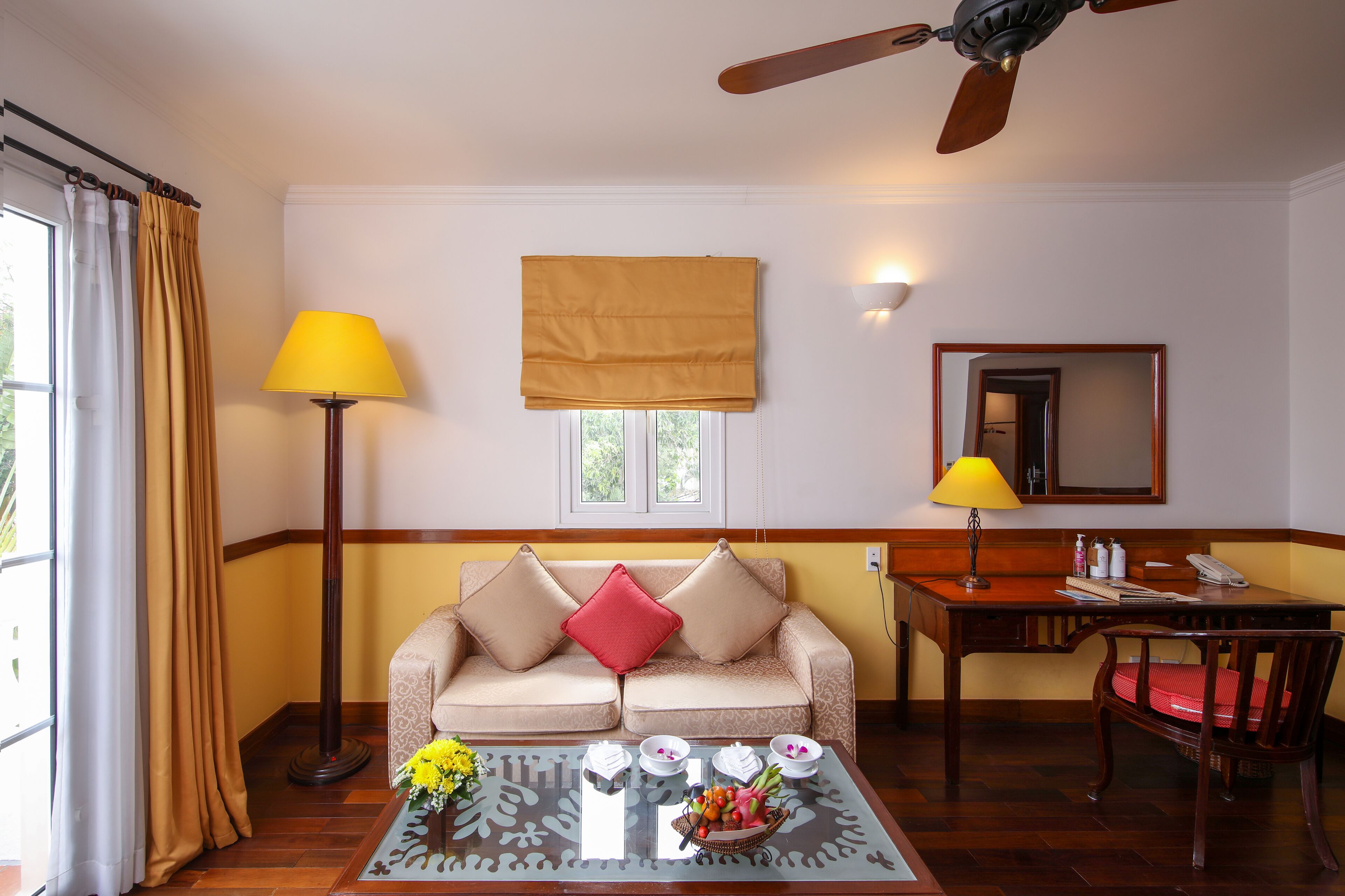 A room with a sofa, a coffee table with flowers, a desk, and a ceiling fan.
