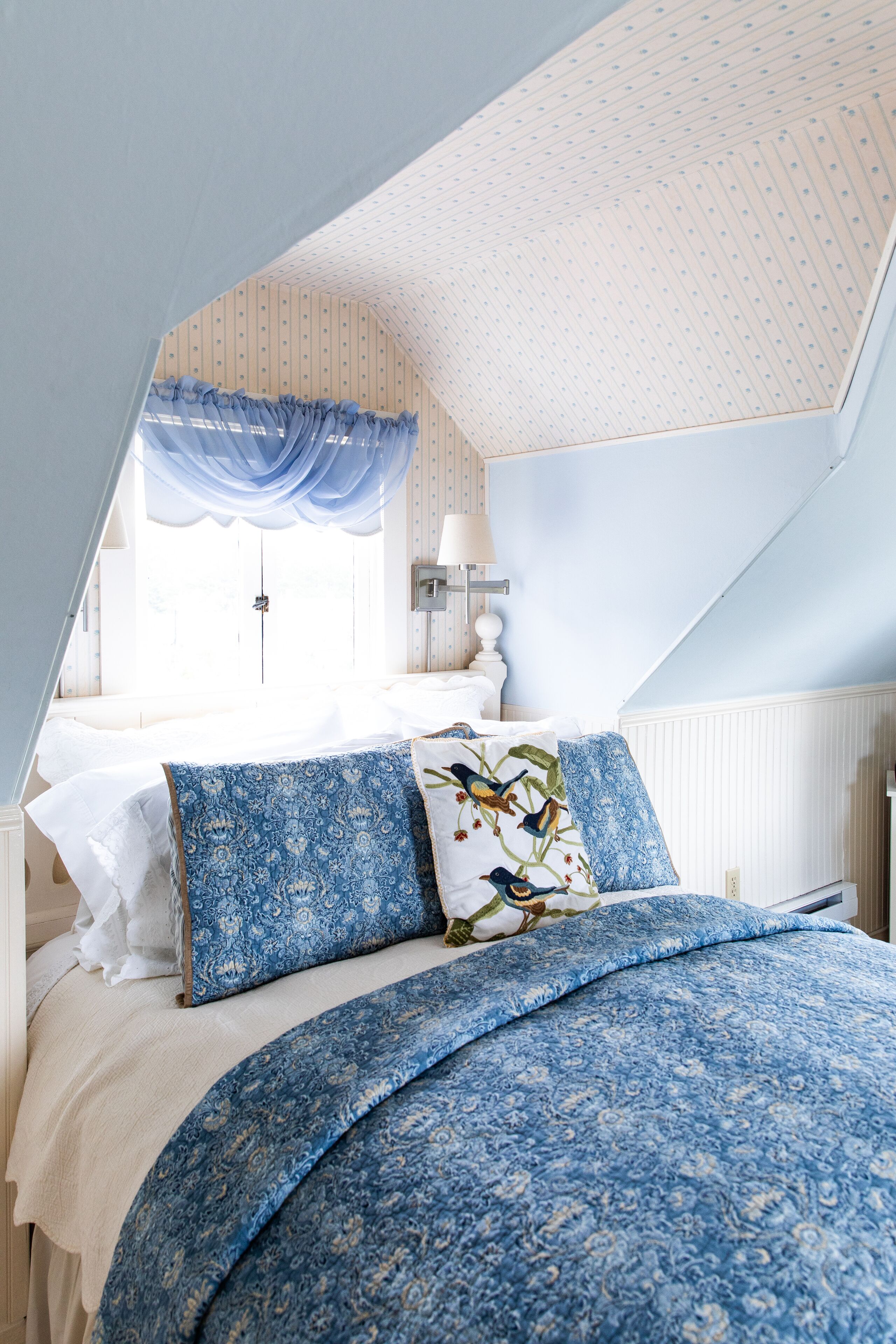 A bedroom with a sloped ceiling, a bed with blue floral bedding, and a window with a blue curtain.