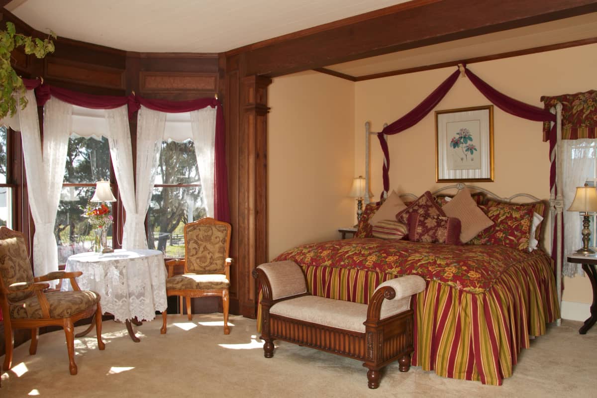 Ein Schlafzimmer mit Himmelbett, zwei Sessel, einem kleinen Tisch und einem Fenster mit Vorhängen.