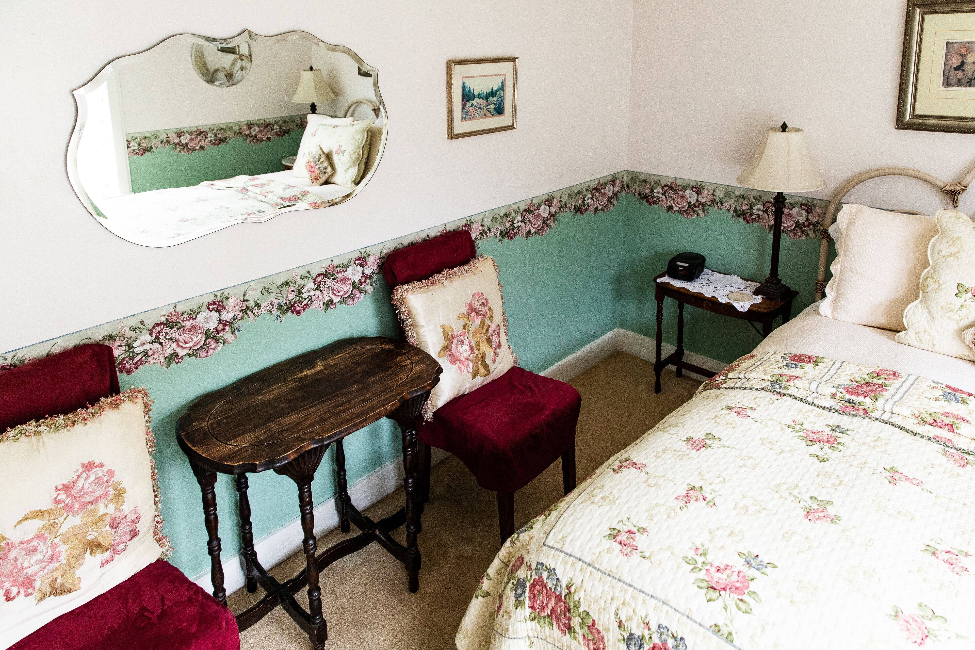 A bedroom with floral wallpaper, a bed with floral bedding, a wooden nightstand, a small table, a chair, and a mirror.