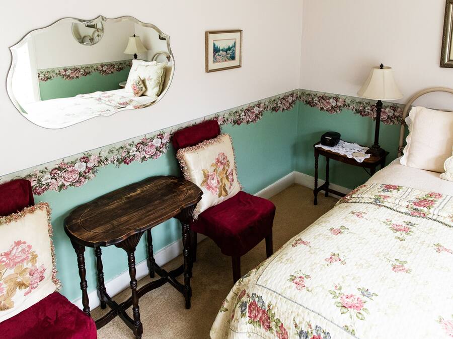 A bedroom with floral wallpaper, a bed with floral bedding, a wooden nightstand, a small table, a chair, and a mirror.