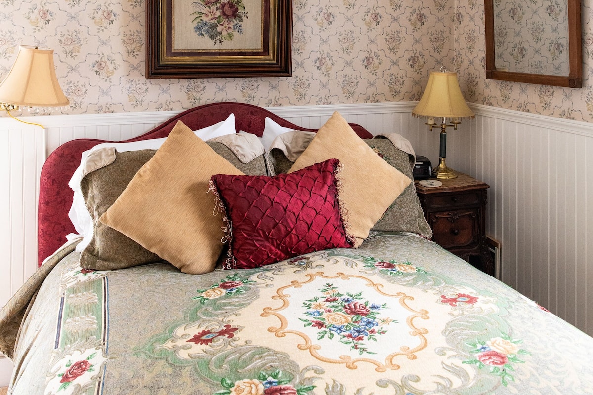 Ein Schlafzimmer mit floraler Tapete, einem Bett mit vielen Kissen, einem Nachttisch mit Lampe und einem gerahmten Bild an der Wand.