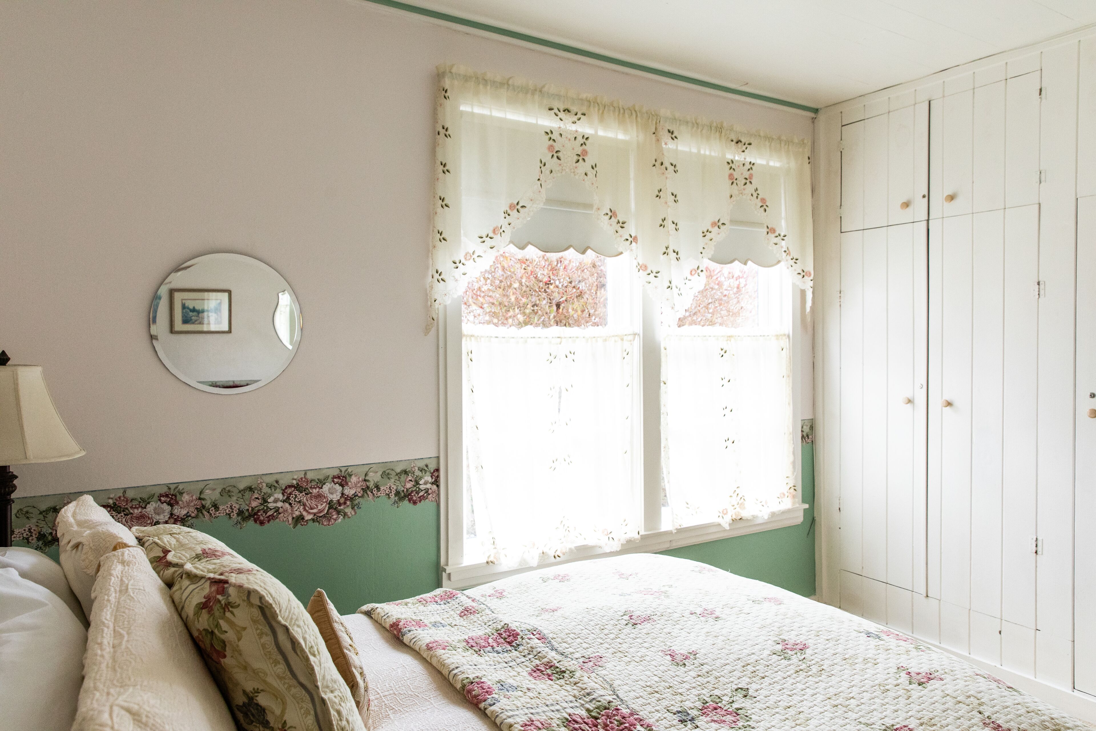 Un dormitorio con cama, ventana con cortinas florales, armario y un espejo redondo en la pared.