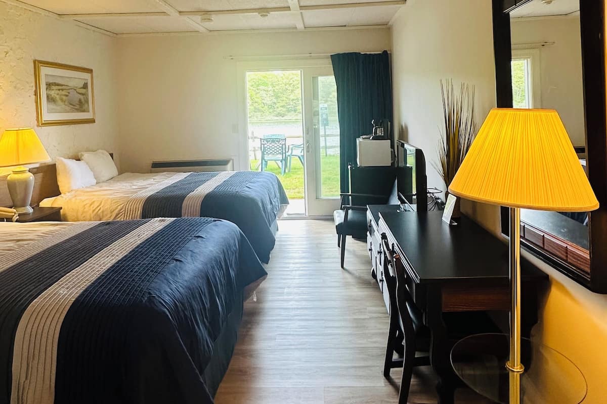A hotel room with two beds, a desk, a chair, a lamp, a mirror, and a view of the outdoors.
