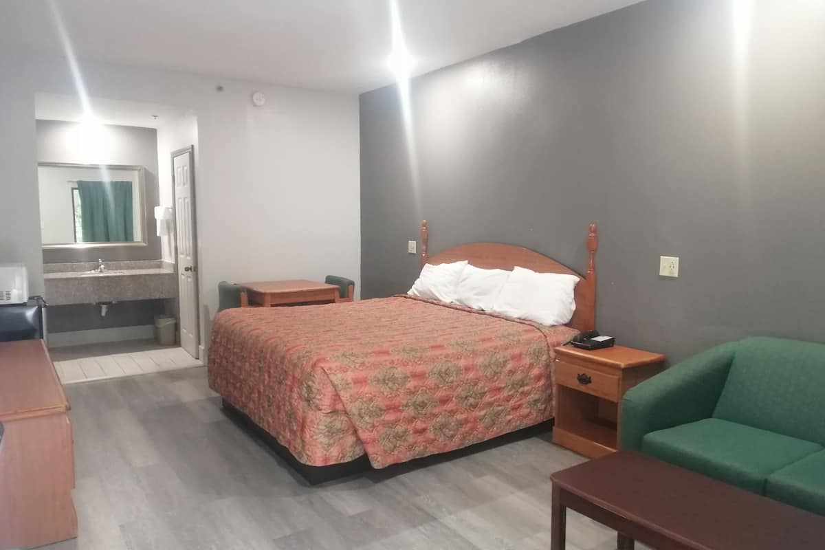 A hotel room with a bed, a green armchair, a wooden nightstand, a small table, a bathroom with a mirror and sink, and a window with curtains.