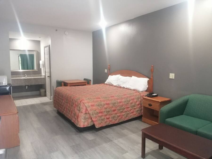 Habitación de hotel con cama, un sillón verde, una mesita de noche de madera, una pequeña mesa, un baño con espejo y lavamanos, y una ventana con cortinas.