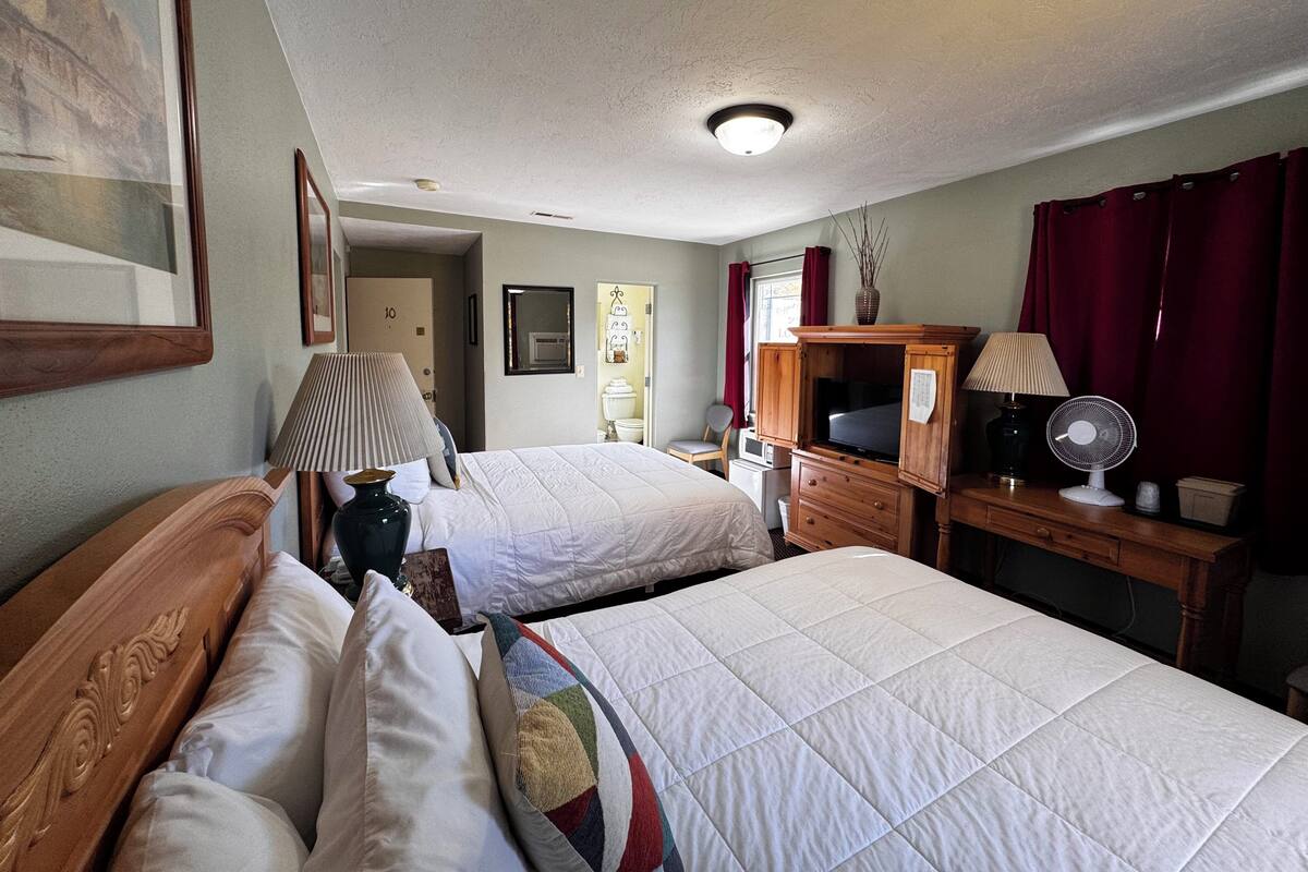 A hotel room with two beds, a desk with a computer, a lamp, and a fan.