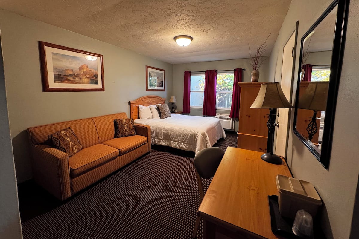 A hotel room with a bed, a wooden desk, a wicker sofa, and two framed pictures on the wall.