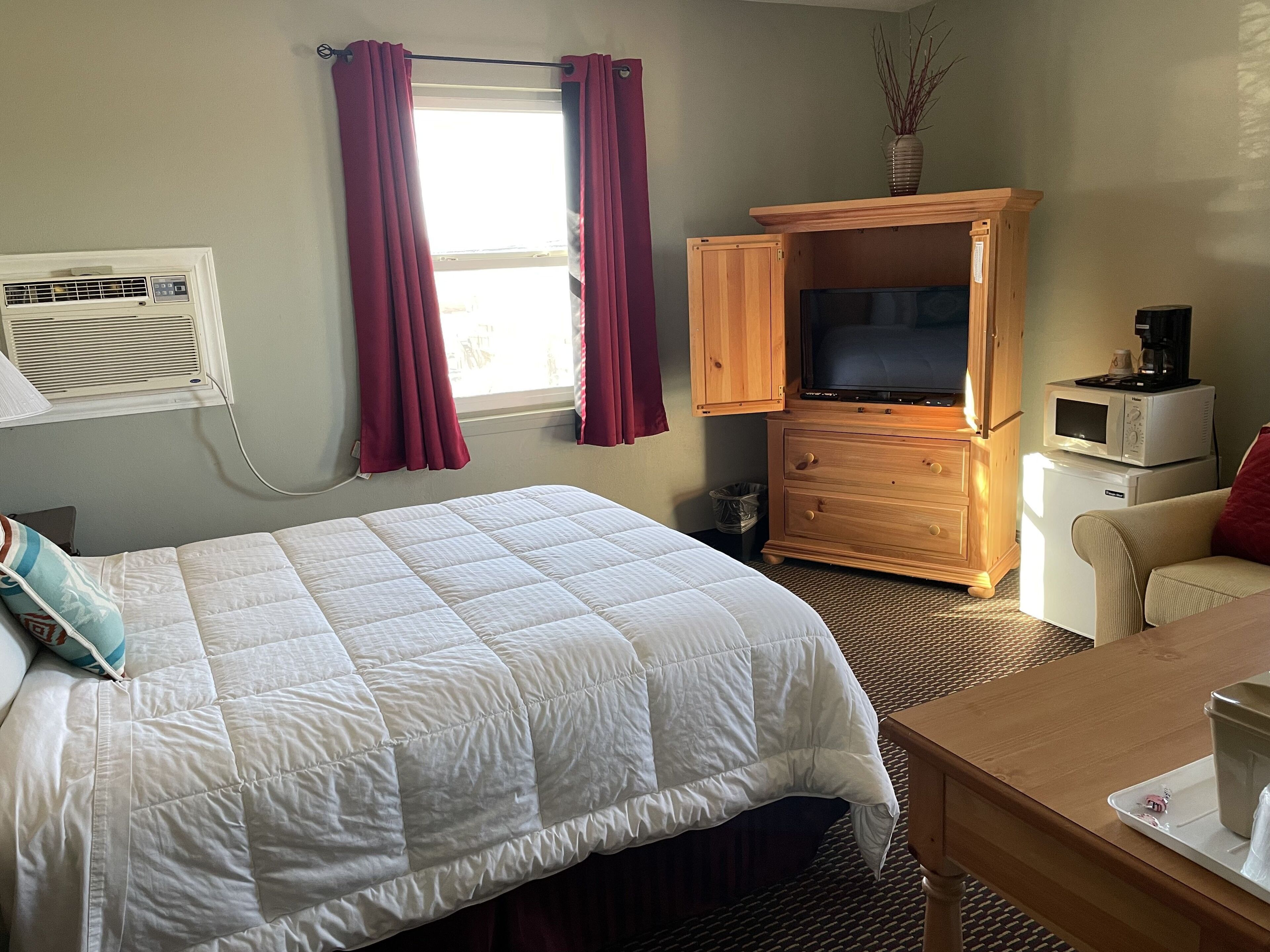 Quarto de hotel com uma cama grande, um móvel de madeira para a TV com uma televisão de tela plana, micro-ondas e uma pequena mesa com uma xícara.