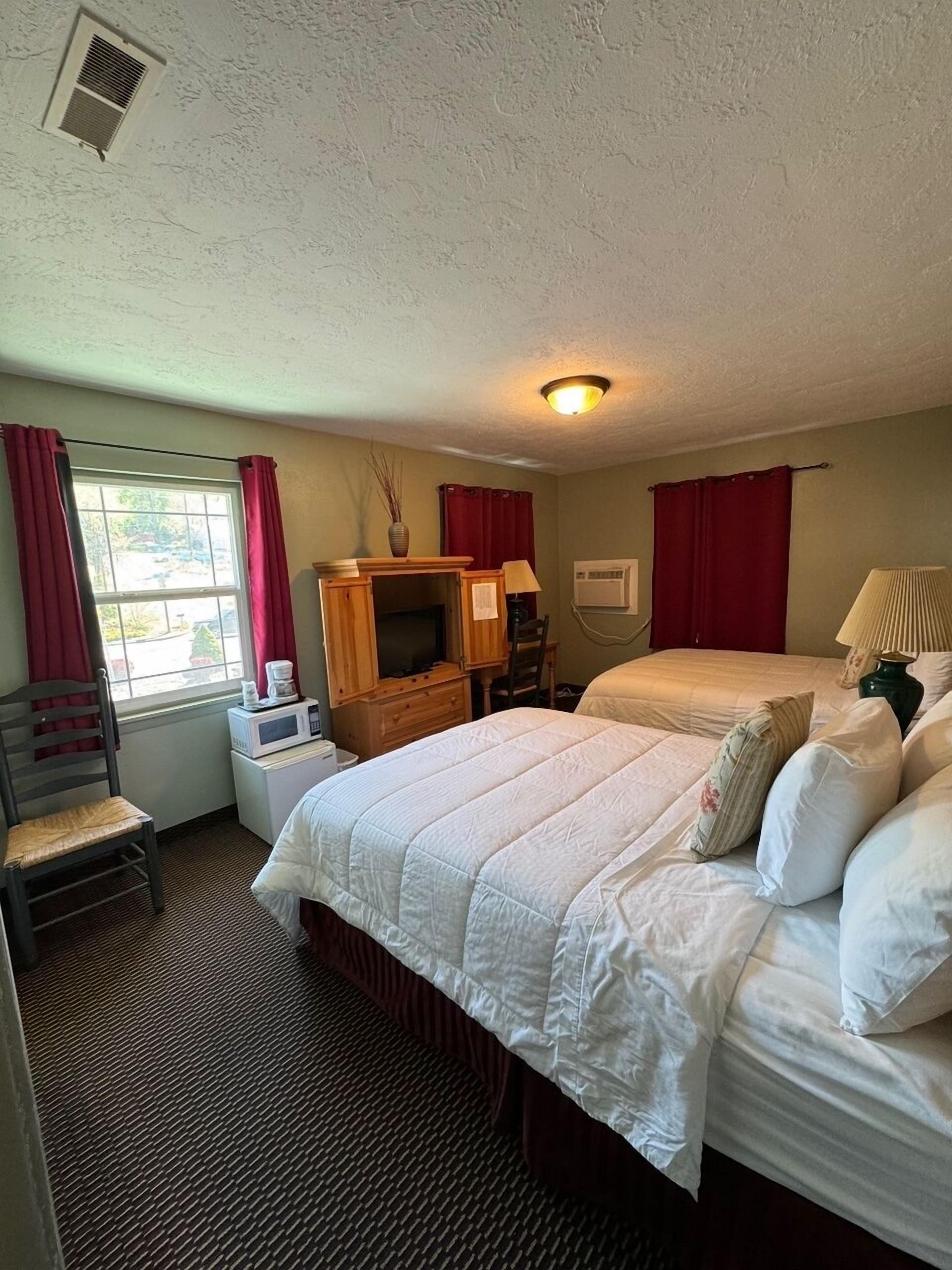 A hotel room with a large bed, a desk with a computer, a chair, a window with curtains, and a ceiling vent.