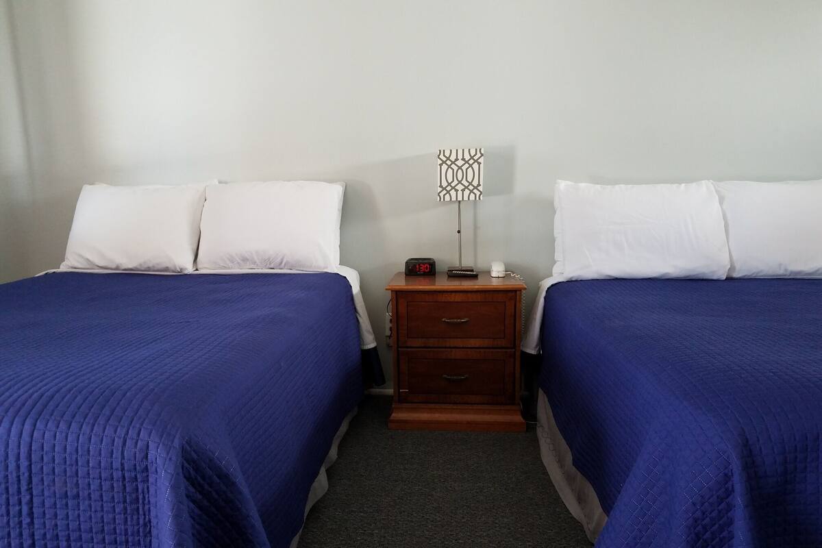 A hotel room with two beds, each with white pillows and blue bedspreads. There is a wooden nightstand with a clock and a phone between the beds.