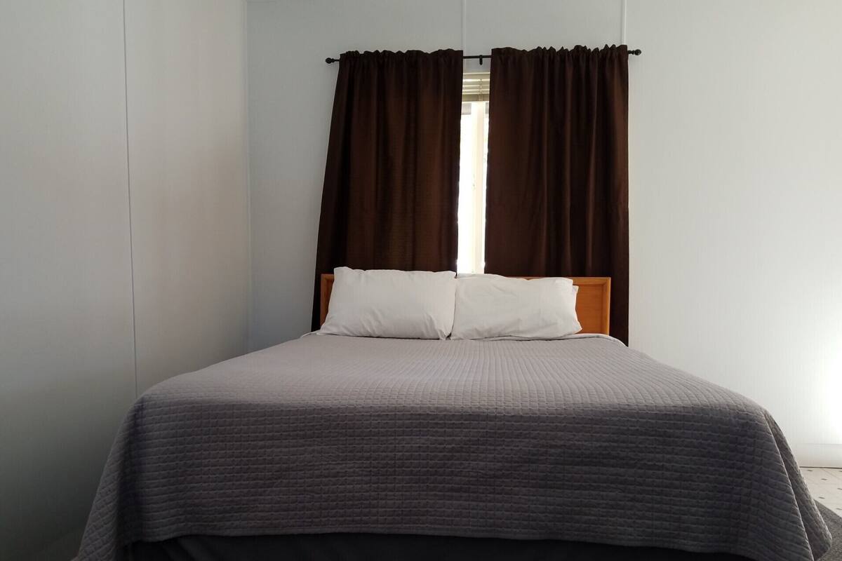 A single bed with a grey quilt, two white pillows, and dark brown curtains.
