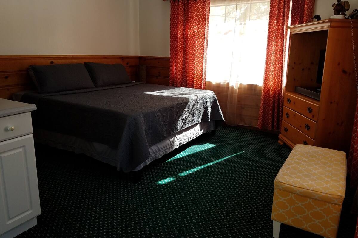 A hotel room with a bed, a wooden dresser, a desk, and a window with curtains.