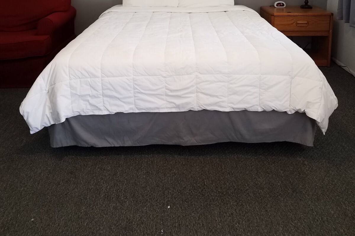 A hotel room with a bed, a red chair, a nightstand with a lamp, and a painting on the wall.