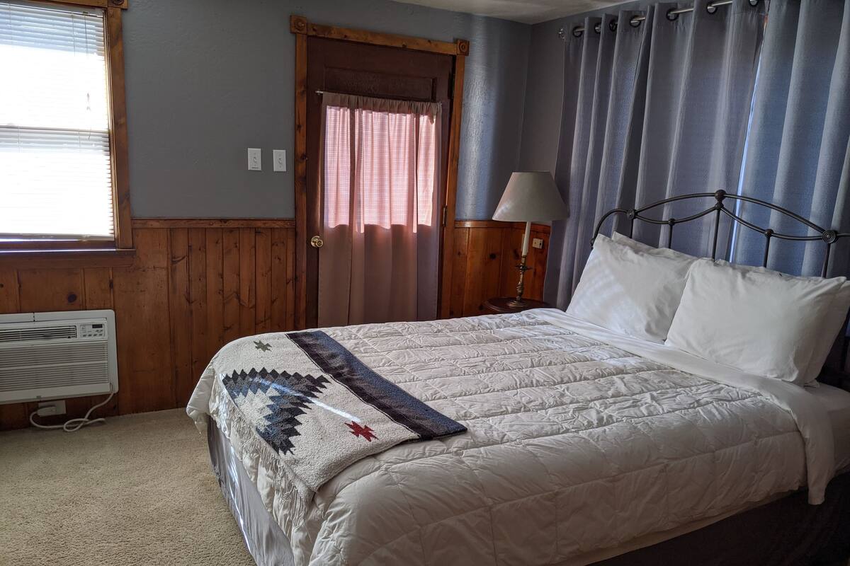 A bedroom with a bed, a nightstand, a lamp, a window with blinds, and a door.