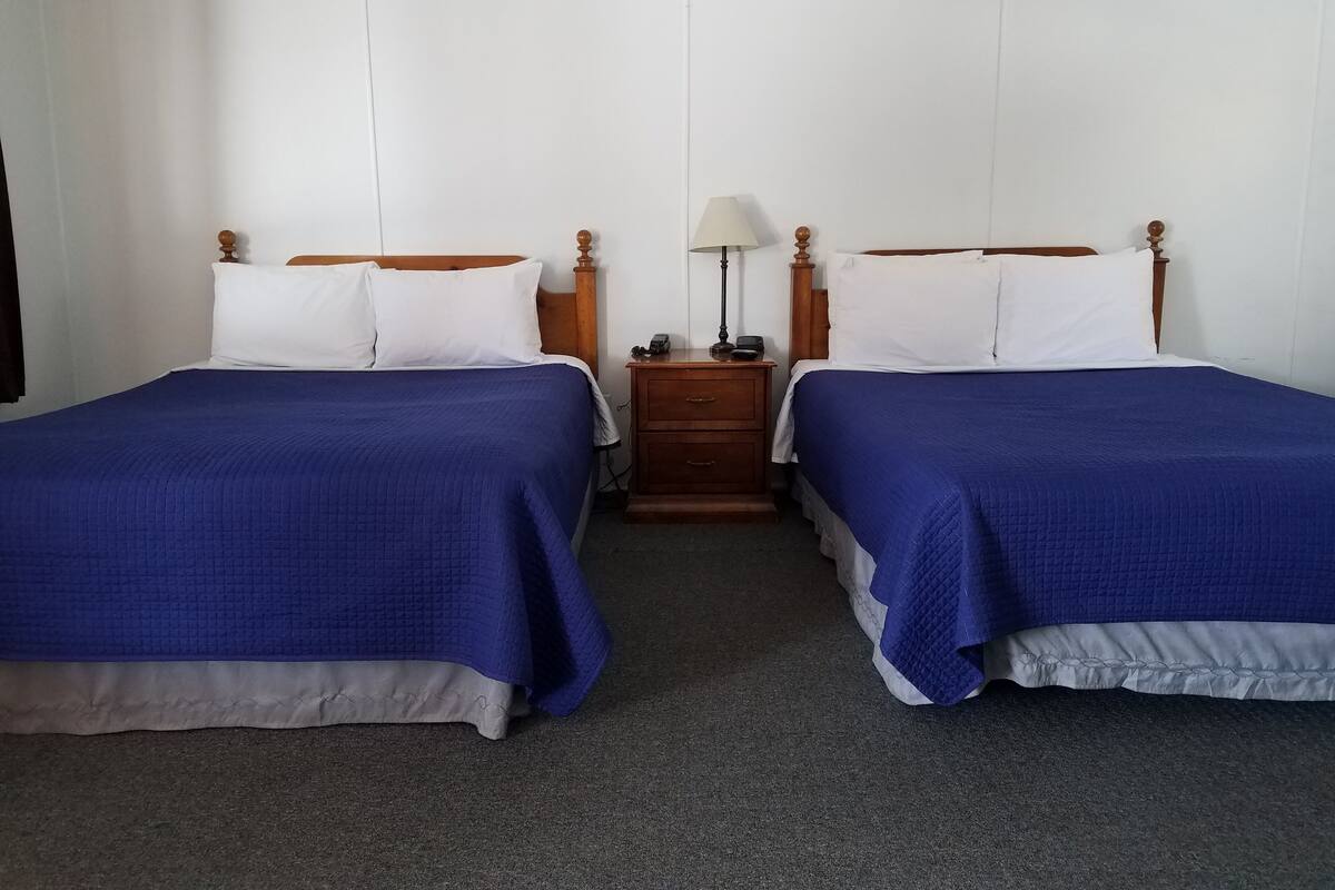 A hotel room with two beds, each with a wooden headboard and white bedding. There is a nightstand with a lamp and a small table between the beds.