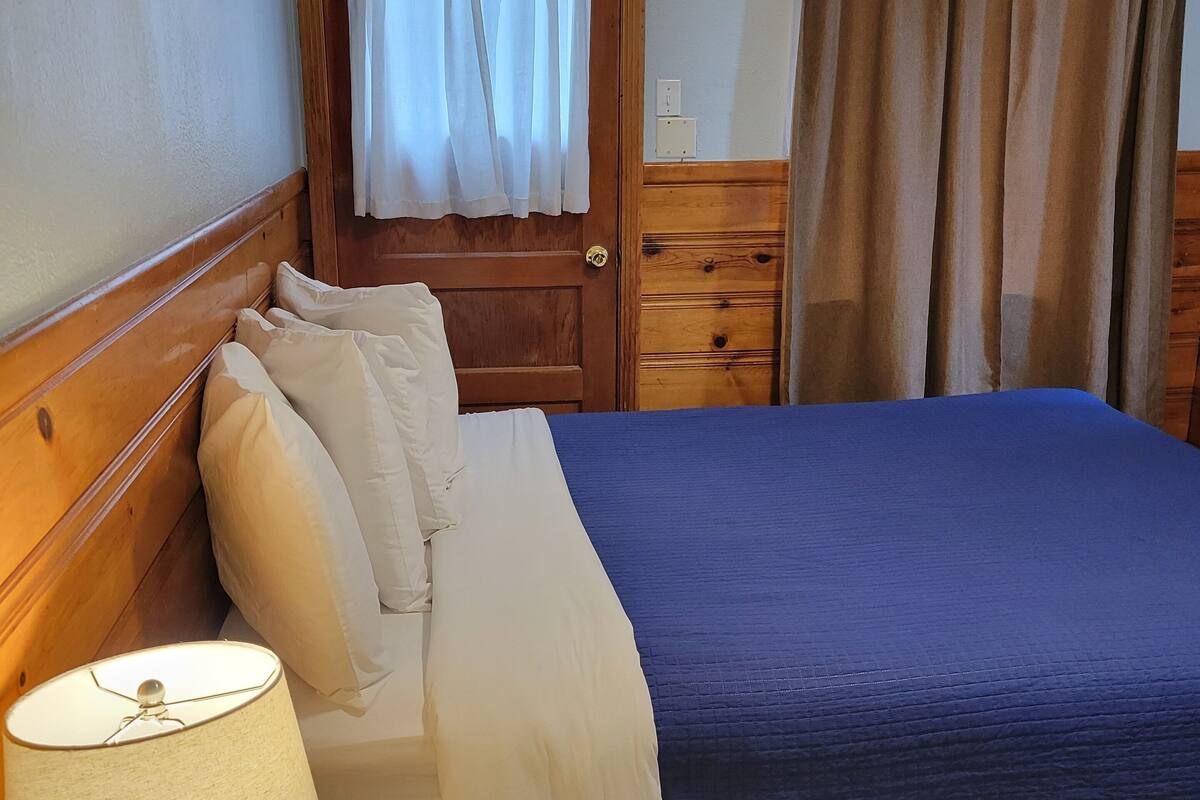 A neatly made bed with a blue bedspread, a wooden headboard, and a bedside table with a lamp.