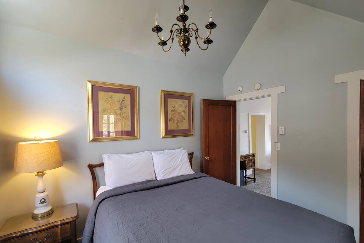 A bedroom with a bed, a nightstand, a lamp, two framed pictures, a chandelier, and a door leading to another room.