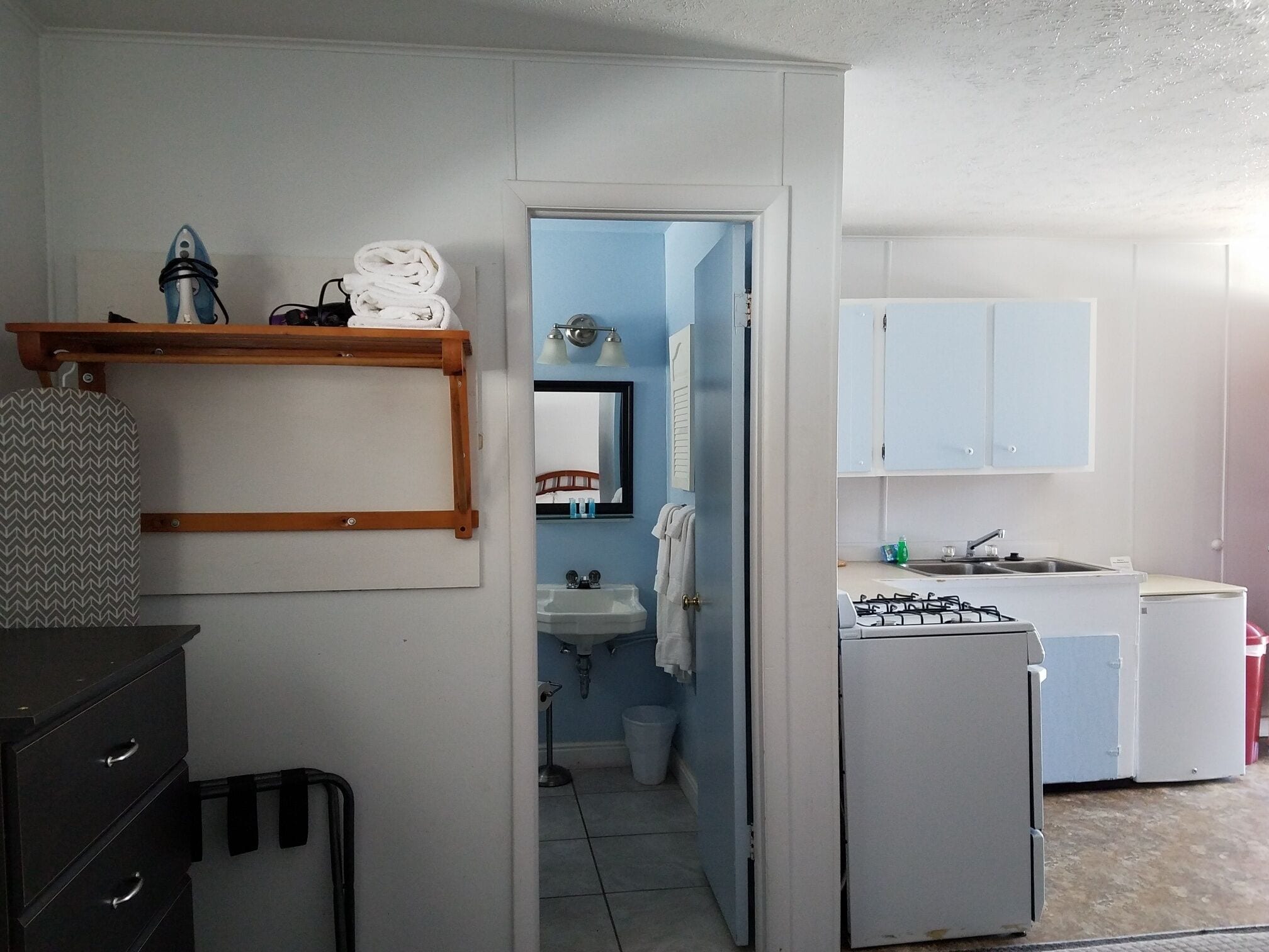 Cocina compacta con estufa a gas, pileta y alacenas. Un vanitory da paso a un baño con un espejo y una toalla colgada en la pared.