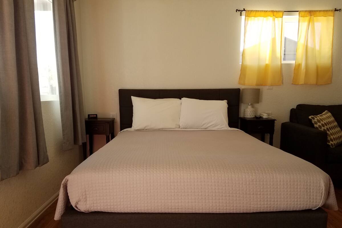 A neatly made bed with a quilted cover, a bedside table with a lamp, a chair with a patterned cushion, and a window with curtains.