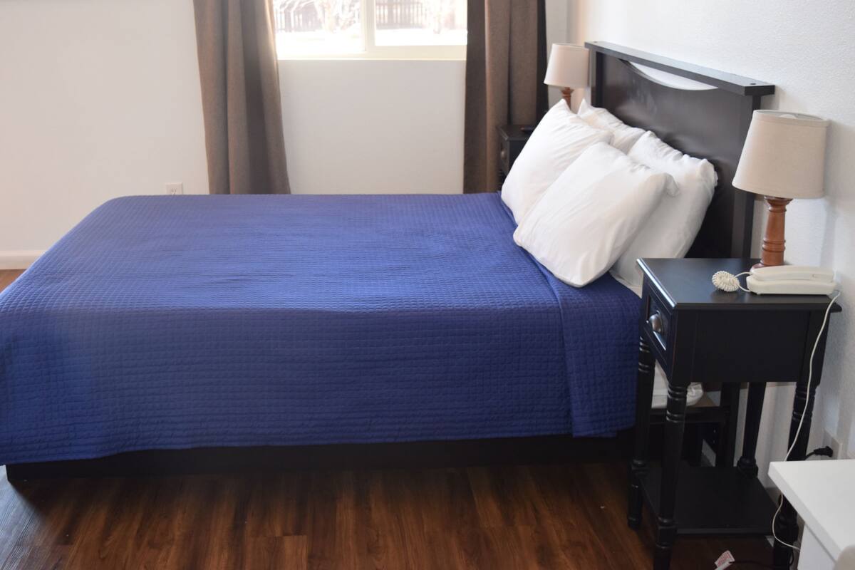 A neatly made bed with a blue bedspread, a wooden headboard, a nightstand with a lamp, and a telephone.