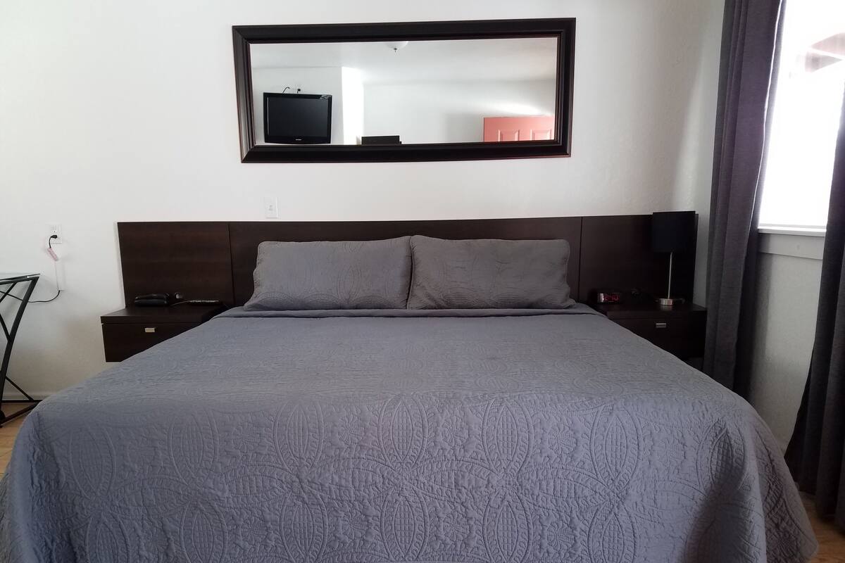 A neatly made bed with a grey patterned bedspread, a dark headboard, and a bedside table with a lamp.