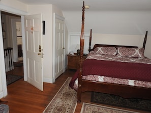A bedroom with a four-poster bed, a wooden floor, a door leading to another room, and a nightstand with a lamp. - Magnolia Manor B&B Inn (Williamsburg)