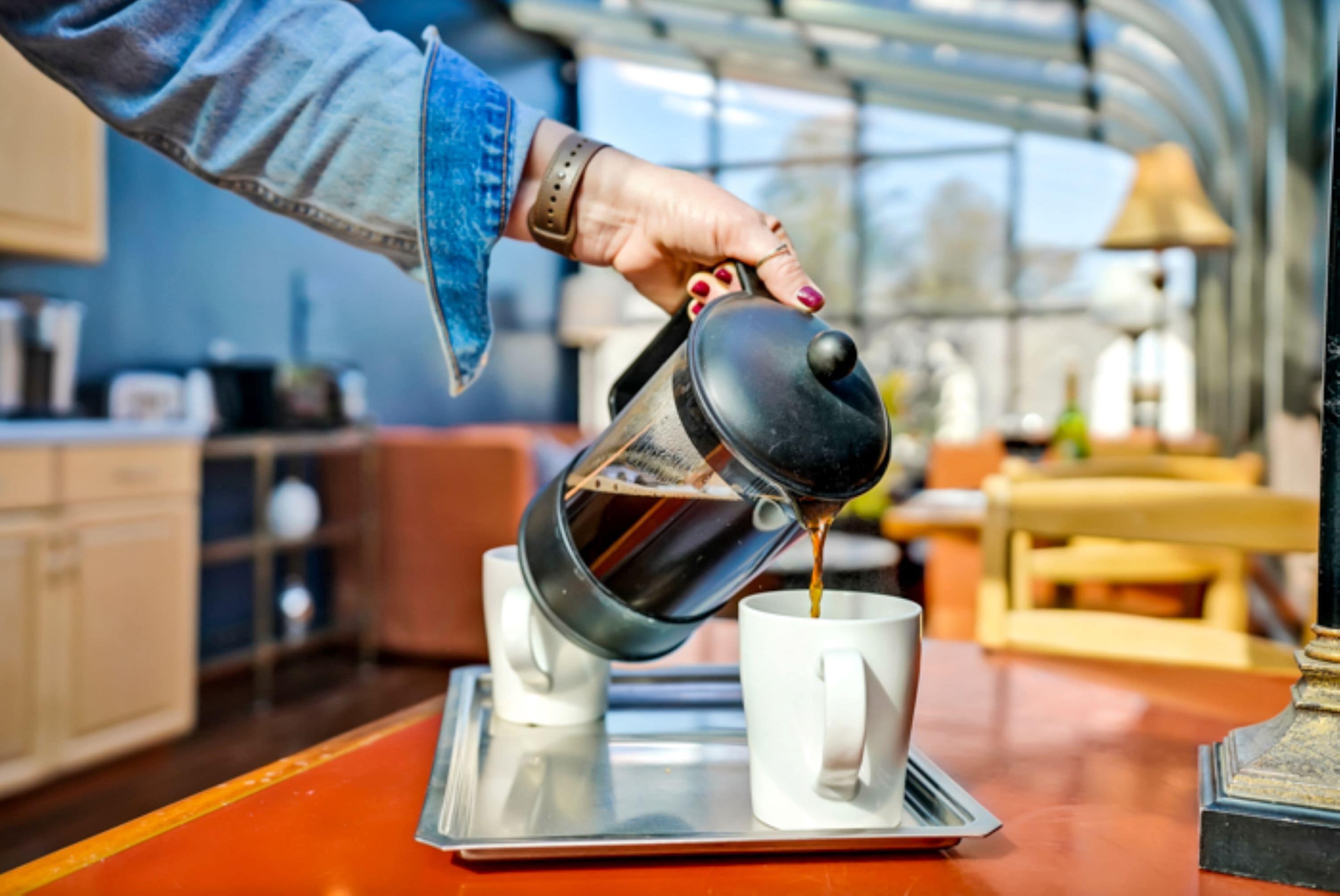 A person pouring coffee into a white mug on a tray in a room with a wooden table and a glass ceiling.