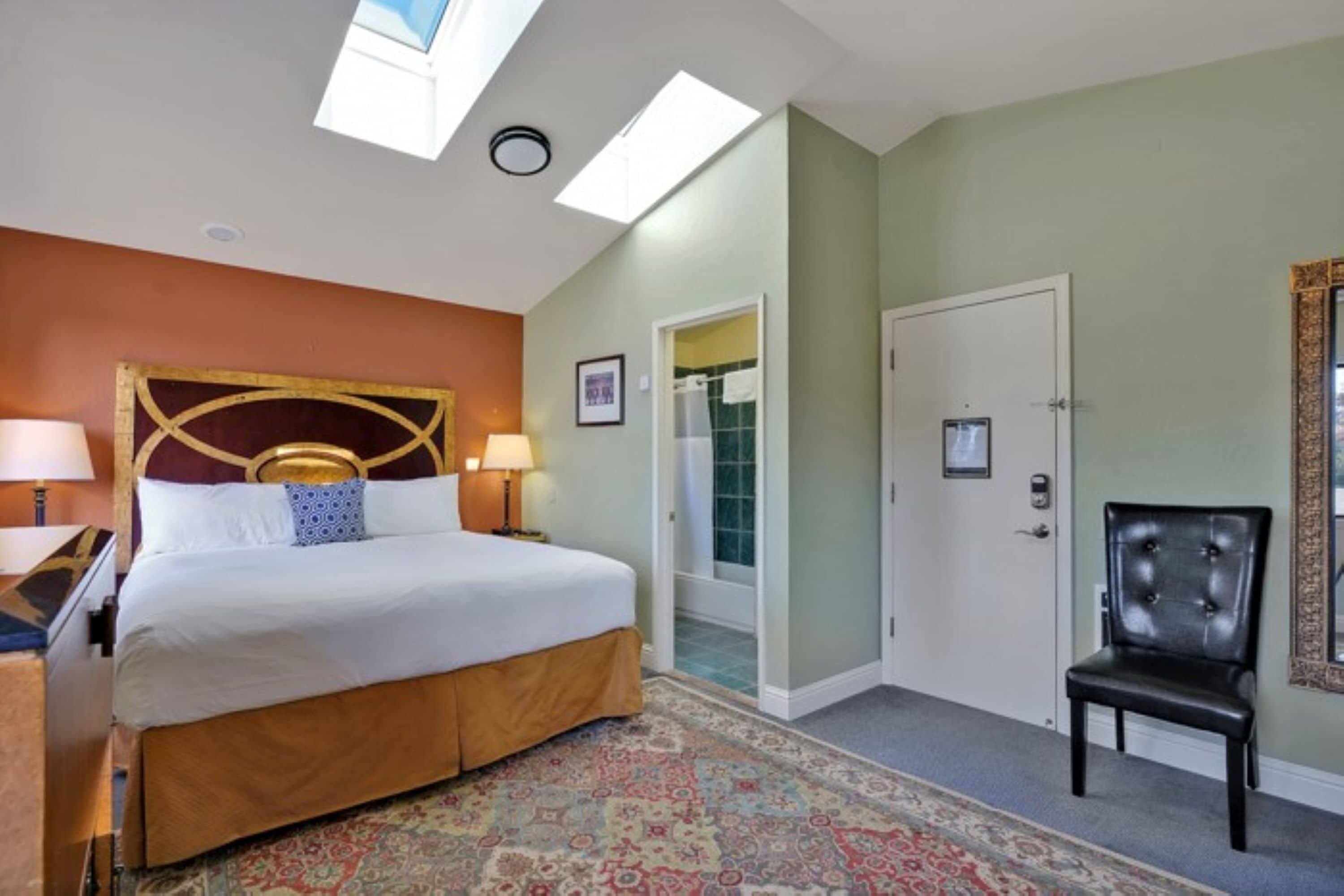 A hotel room with a large bed, a sitting area with a chair, a small table, a nightstand, a window with a view, and a door leading to a bathroom.