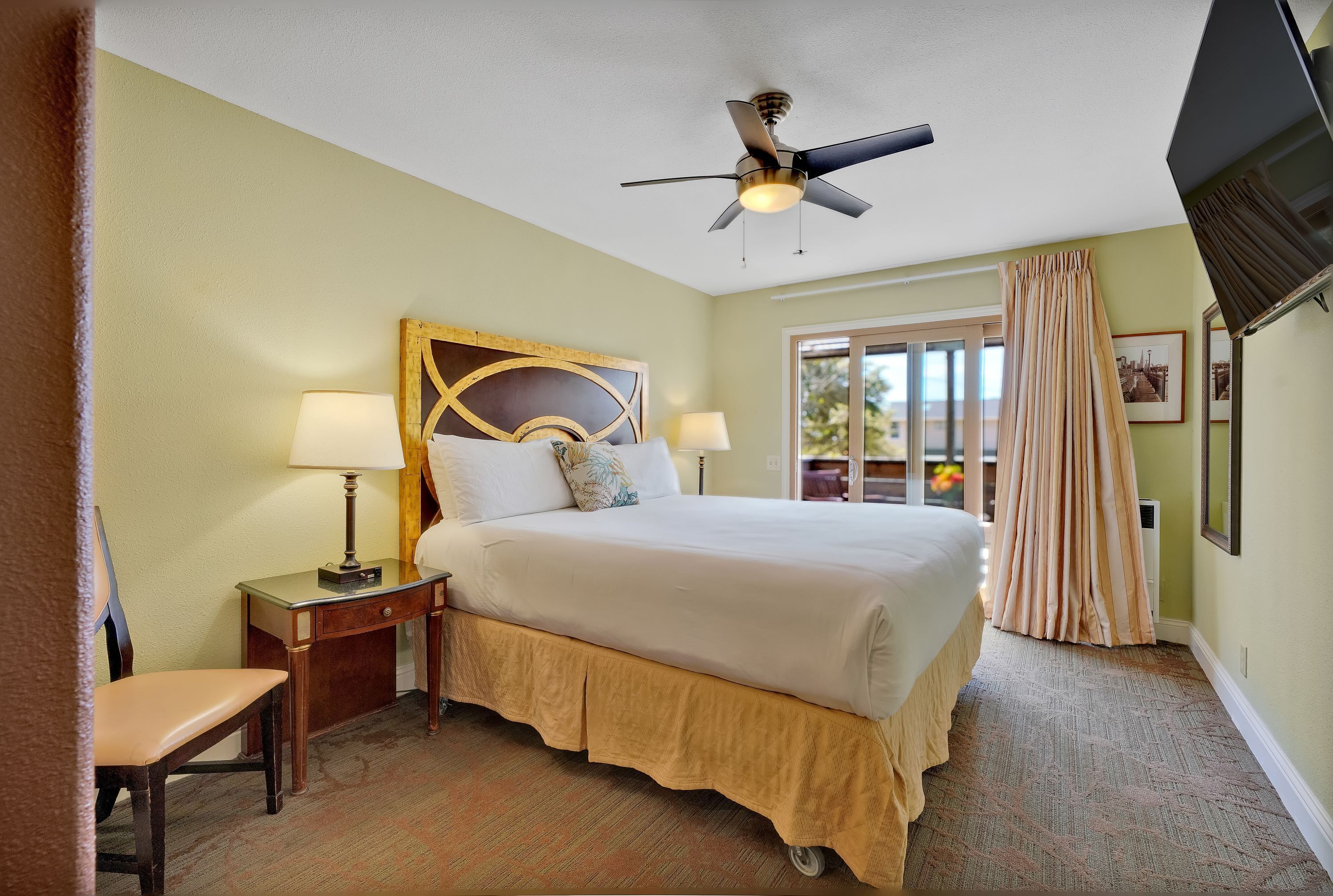 A hotel room with a bed, two bedside tables with lamps, a ceiling fan, a window with curtains, and a view of greenery outside.