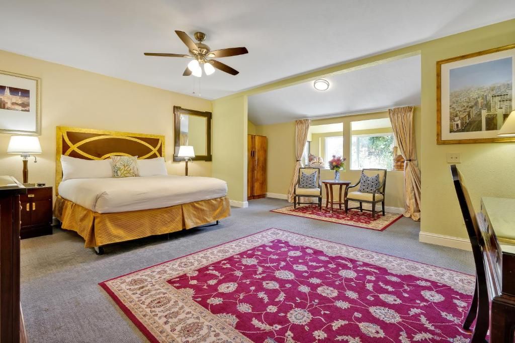 Habitación de hotel con cama, ventilador de techo, ventana con cortinas, una mesita con sillas y una alfombra grande.