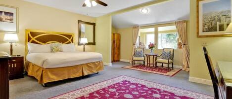 A hotel room with a bed, a ceiling fan, a window with curtains, a small table with chairs, and a large rug.