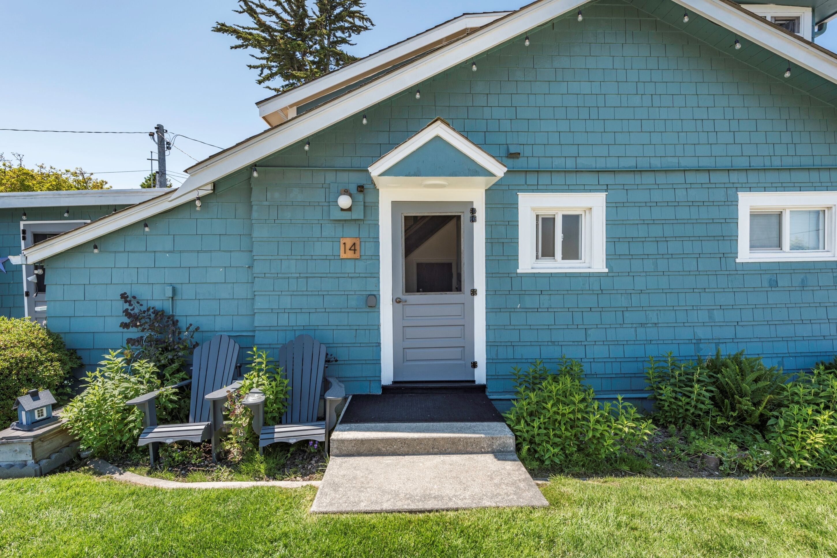 Ein blaues Haus mit weißer Tür, Hausnummer 14, und einer kleinen Veranda mit Adirondack-Stühlen.