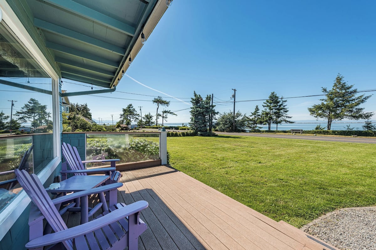 A deck with Adirondack chairs and a glass railing overlooking a grassy area and a road.