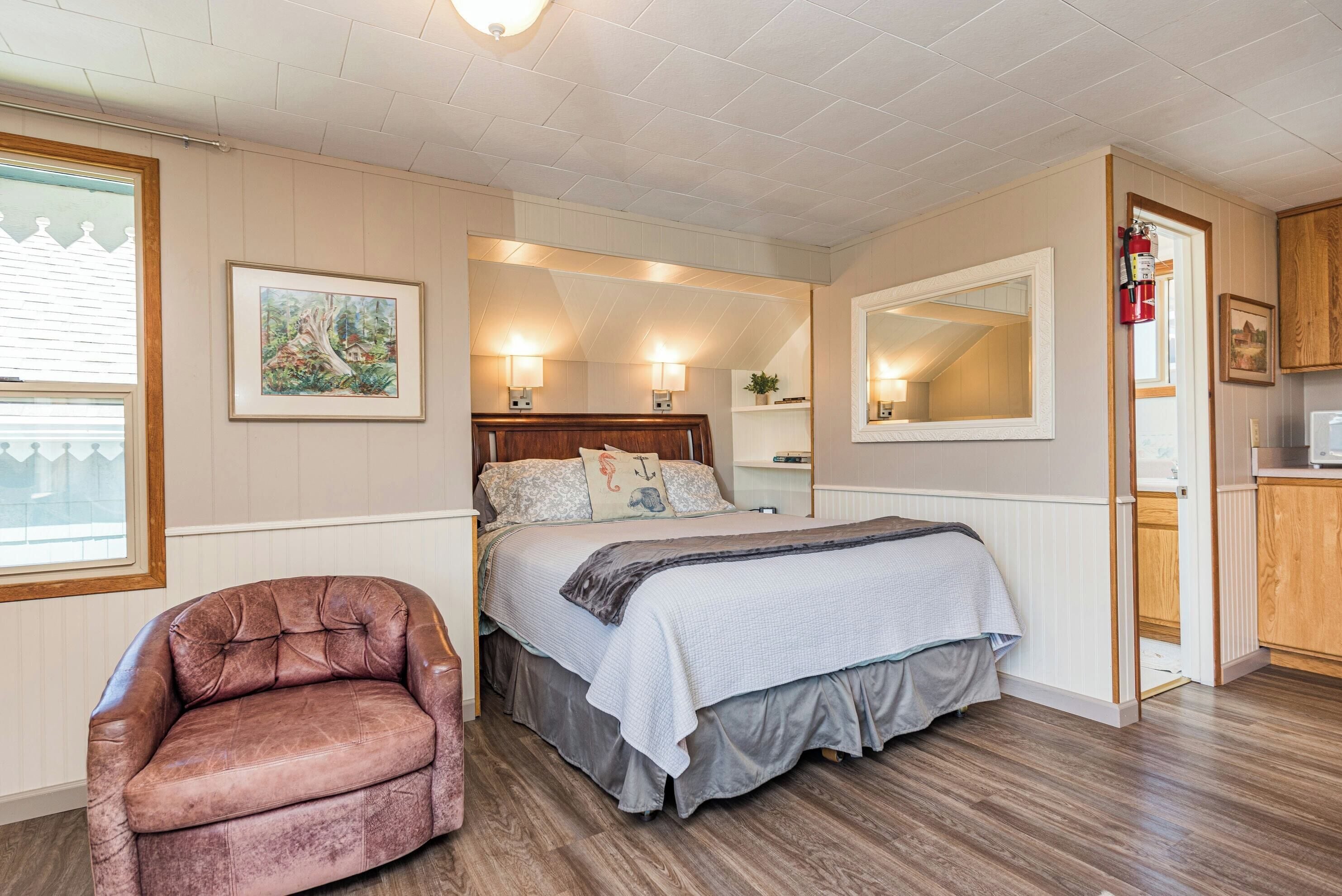A bedroom with a bed, bedside lamps, a chair, a mirror, and a door leading to a kitchen.