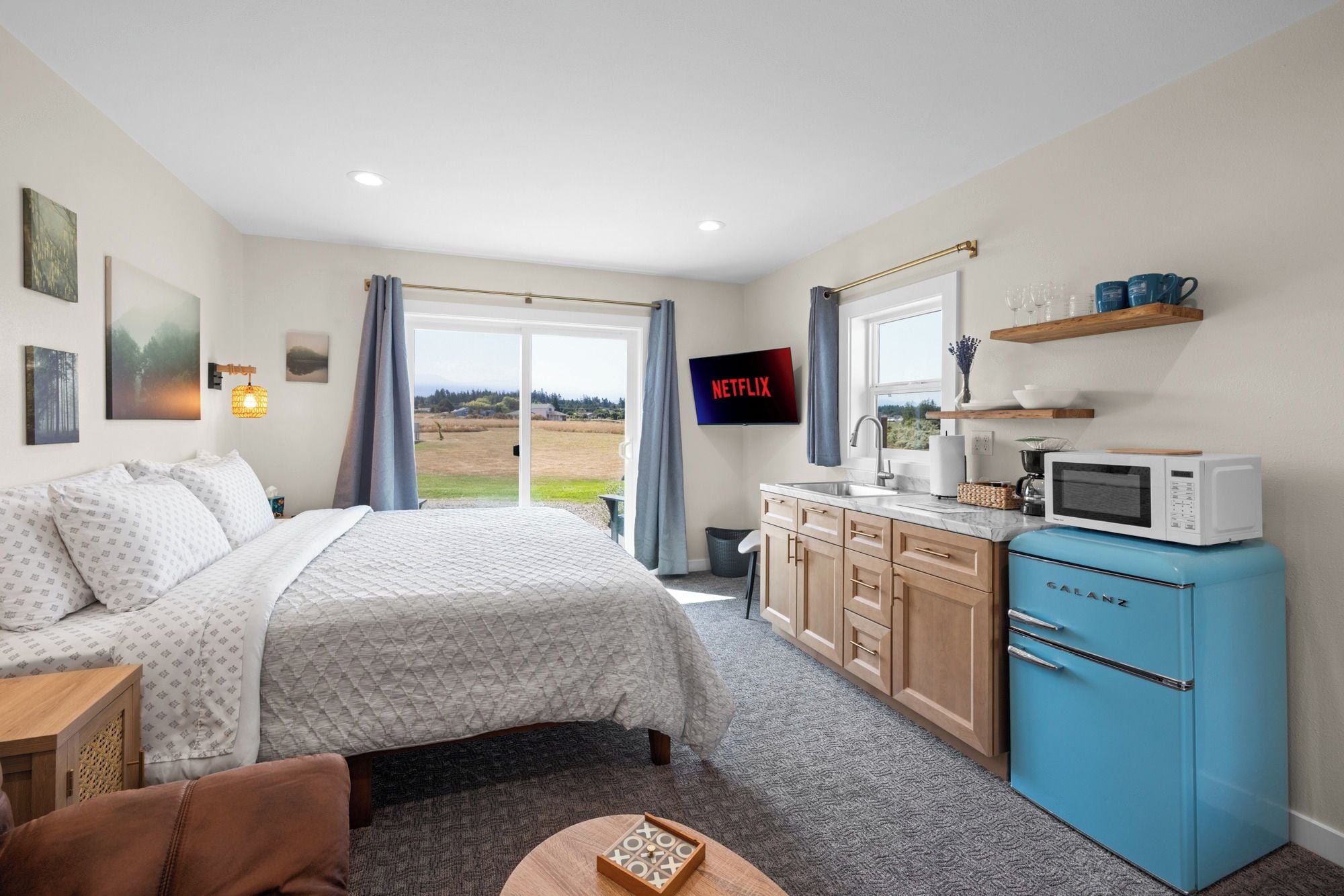 A bedroom with a bed, a desk, a blue refrigerator, a television, and a view of the outdoors.