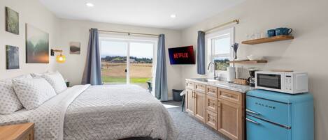A bedroom with a bed, a desk, a blue refrigerator, a television, and a view of the outdoors.
