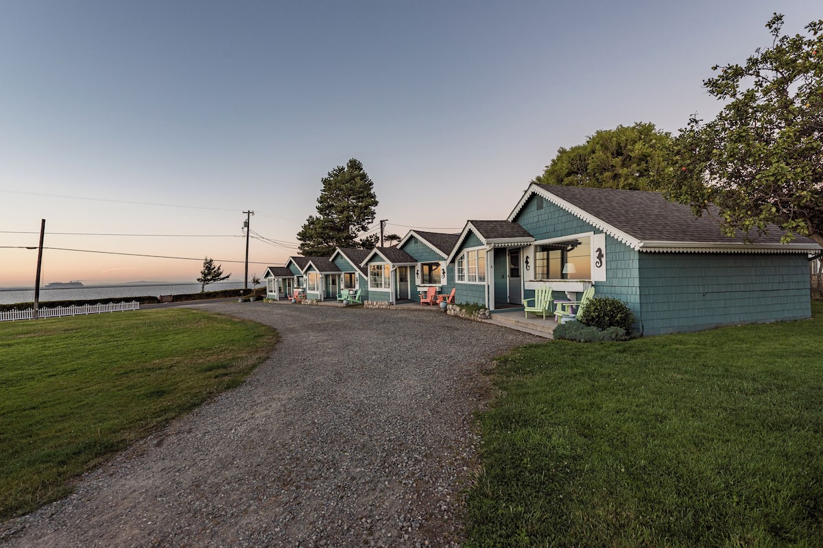 A row of small, single-story buildings with a gravel driveway leading to a grassy area.