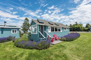 A two-story house with a blue exterior, a front porch with red Adirondack chairs, and a small garden with purple flowers. - Juan de Fuca Waterfront Hotel & Cottages (Sequim)